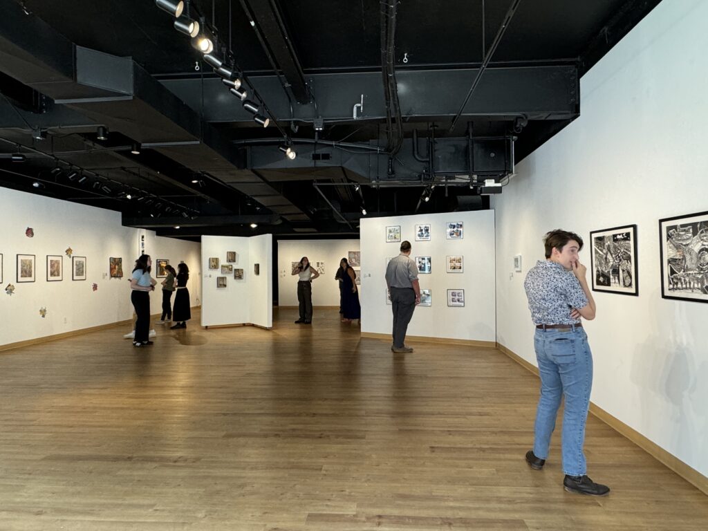 A gallery space with art on the wall. On the right in the foreground is a young person in blue jeans and a grey shirt looking at art. In the background are more people looking at art.