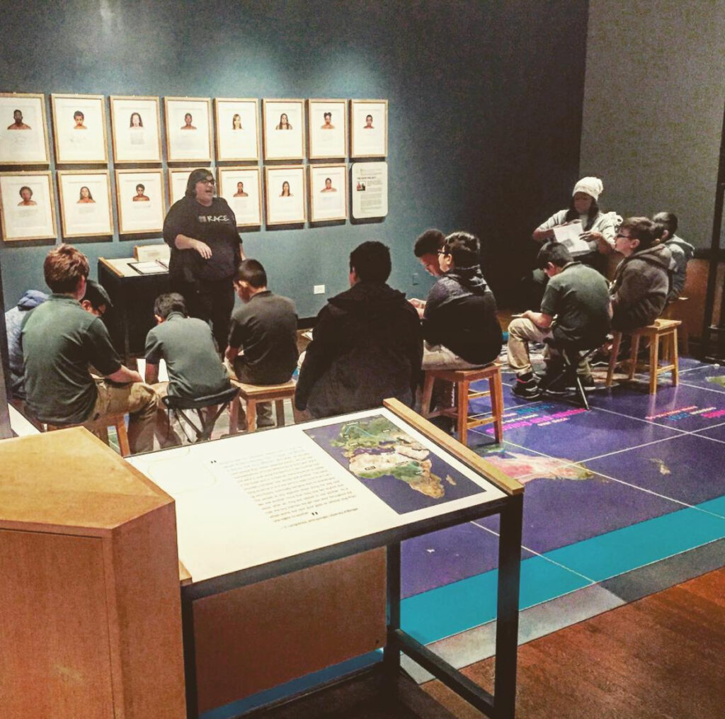 A museum gallery where the walls are green and white and the floor has a map of the world on top of the map are 12 wooden chairs with 12 children the ages of 10 and 11 in green and khaki school uniform looking up at a presenter, dressed in all black standing in front of a display that has various images of people from the shoulder up. 