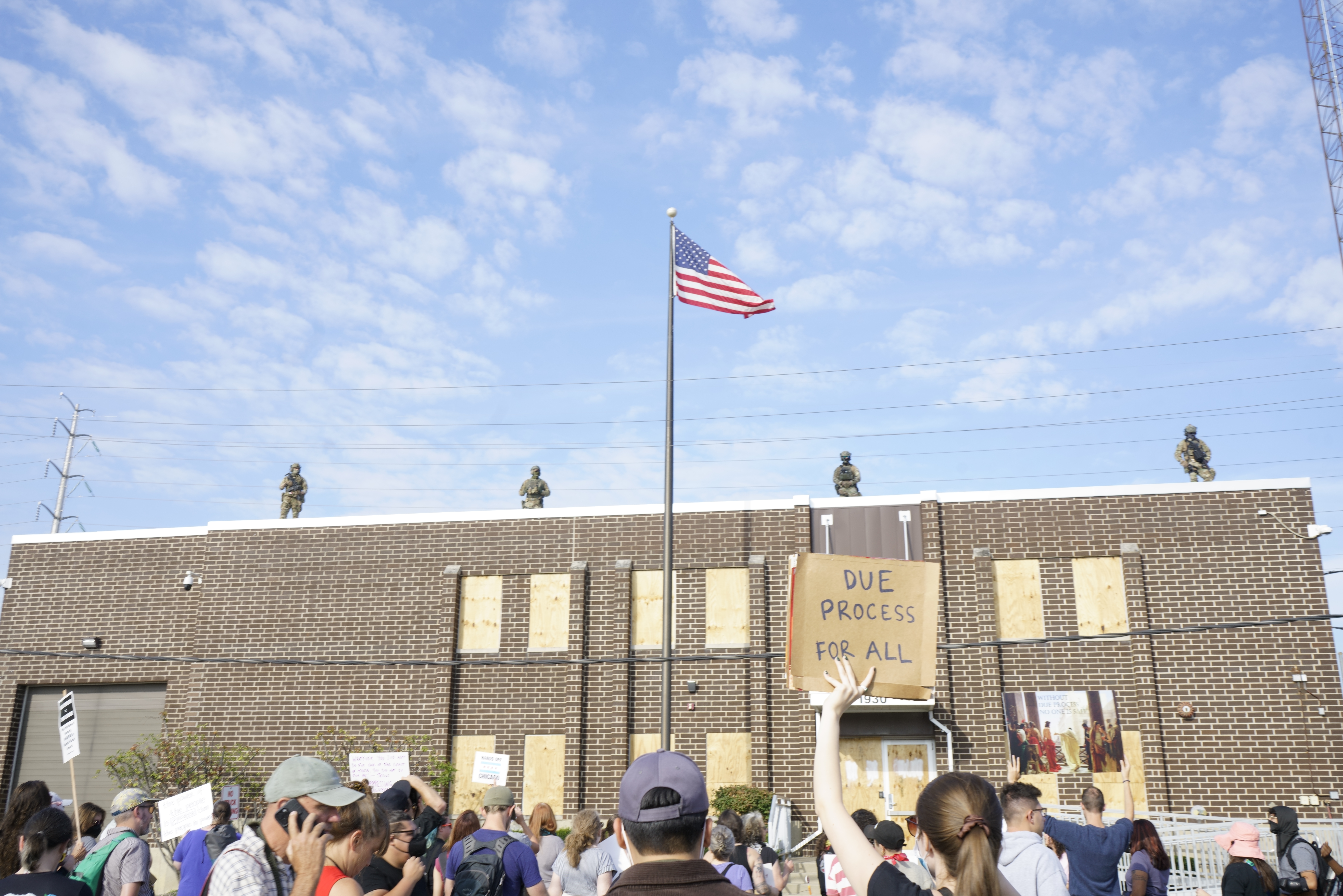 The image shows a blue sky with four armed officers in army uniform, standing on top of a brick building with boarded up windows. The bottom of the image shows the back of protestors heads with one woman holding a cardboard protest sign in her left hand that reads “Due process for all”. A flag pole waving the U.S. flag cuts through the center of the image.