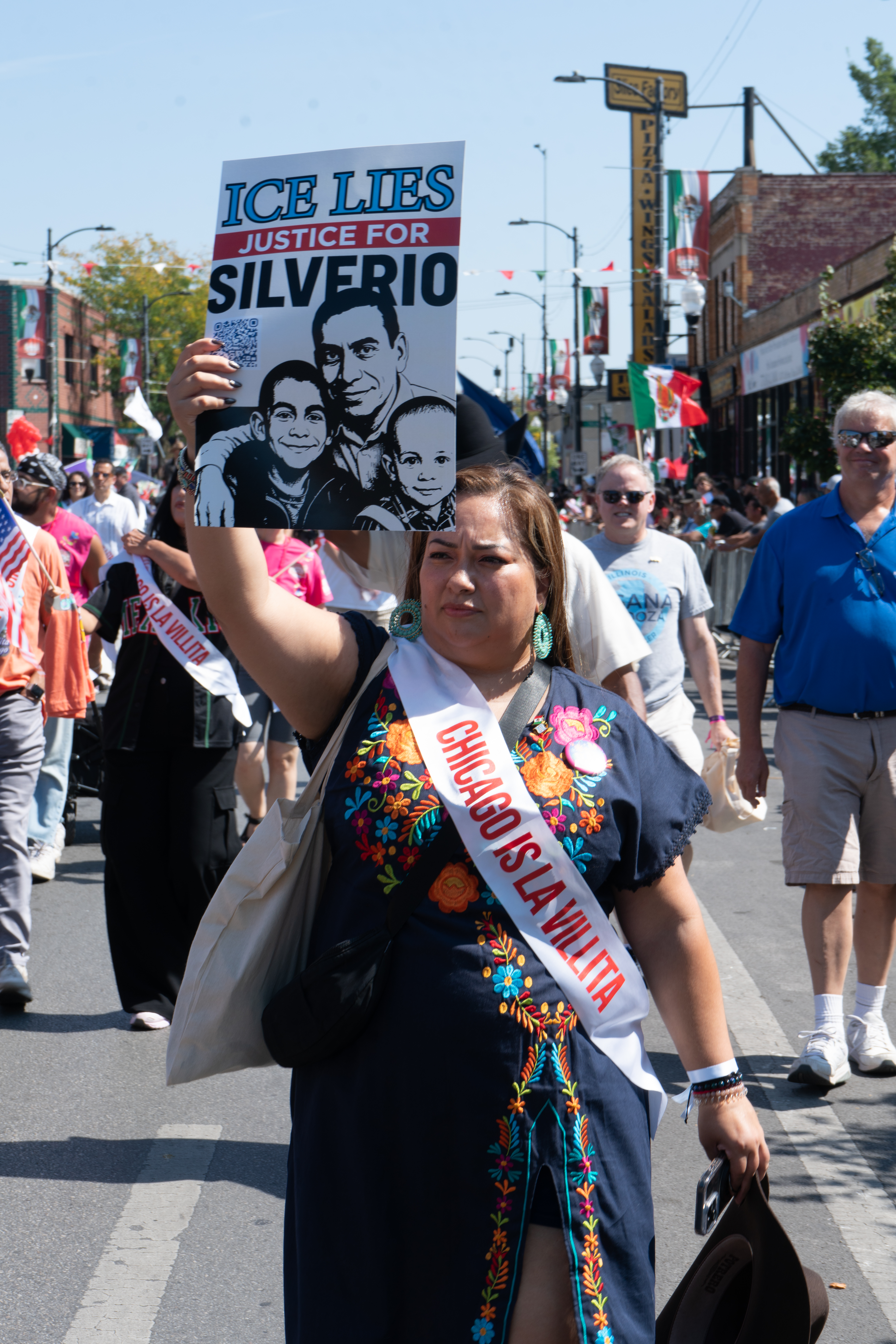 A woman stands in the center wearing a black dress with colorful embroidery and a white sash. In red letters, the white sash reads “Chicago is La Villita”. She is holding a protest sign in her right hand that shows a man’s face along with two children next to him. The sign reads “Ice Lies Justice for Silverio”. In the background, a group of people are walking down the street holding U.S. and Mexican flags.