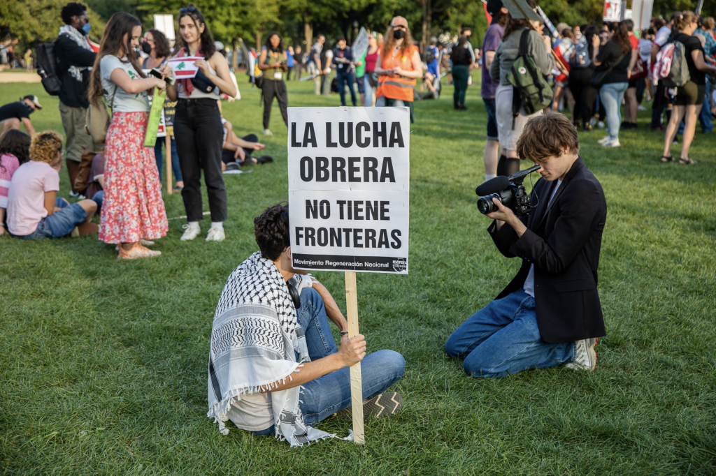 ○ People gather in a grassy field. The background shows multiple groups of people standing and sitting down. The foreground shows a male sitting down, wearing a Palestinian keffiyah and jeans, and holding a white sign with black letters that reads “La Lucha Obrera No Tiene Fronteras” translating to “The Worker’s Struggles Know No Borders”. To the right of him there is a male photographer kneeling and pointing a video camera at the protestor.