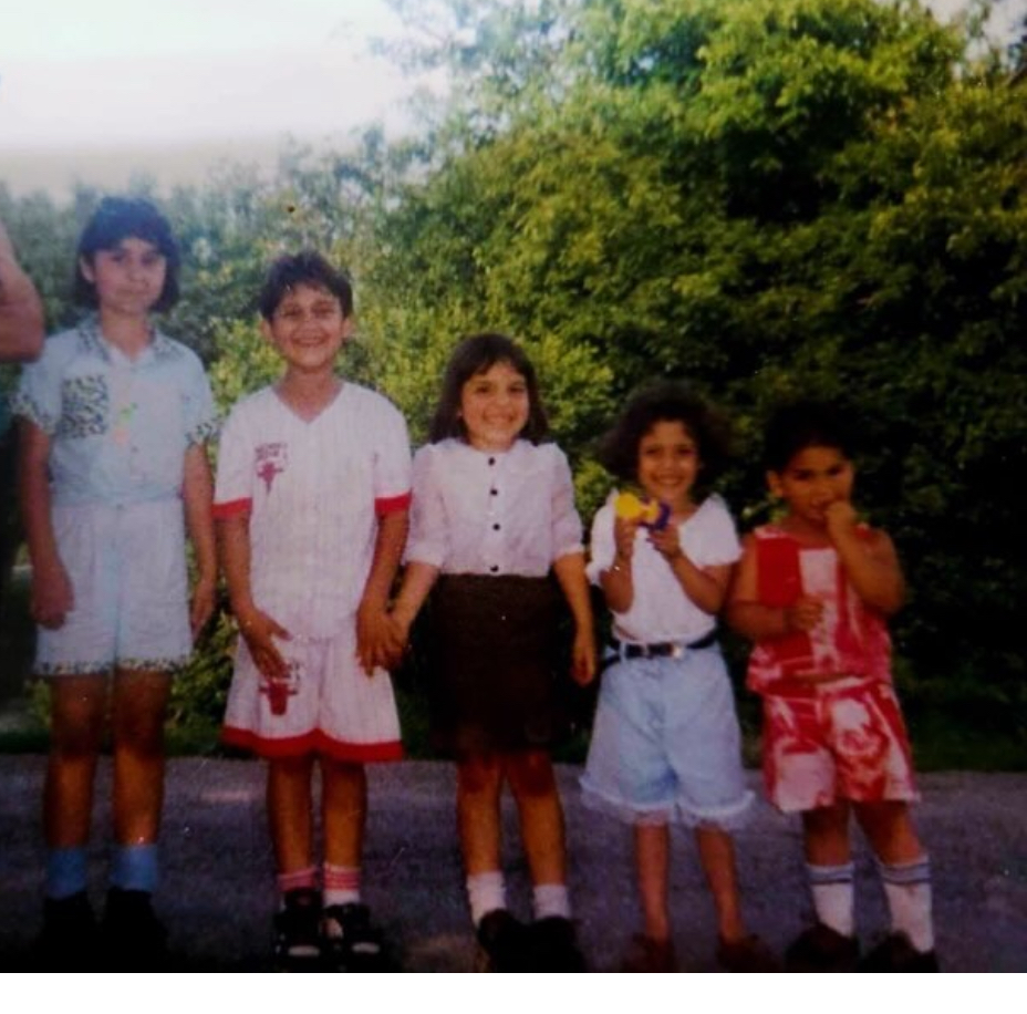 Five Mexican children standing in front of a green bush from tallest to shortest. From let to right: a 12-year-old girl wearing a light blue shirt and shorts with a short bob looks straight at the camera. A 10-year-old boy wearing a white shirt and shorts with a red bull logo of the Chicago Bulls basketball team, smiling, staring at the camera and holding hands with a little girl to his right. next to the boy is an eight-year-old girl wearing a white shirt button up with a green skirt, smiling at the camera with shoulder length hair. Next to her is another little girl with a curly bob a white shirt, jean shorts holding on to something yellow, smiling. Next to her is a little boy with a shirt and shorts that are red and white. He has his hands in his mouth like he’s eating something and he’s staring at the camera.