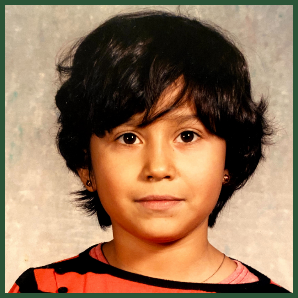 a little Mexican girl with a very short haircut, little round golden earrings, a black and red striped sweater, looking directly at the camera. The background is a light and dark brown backdrop. 