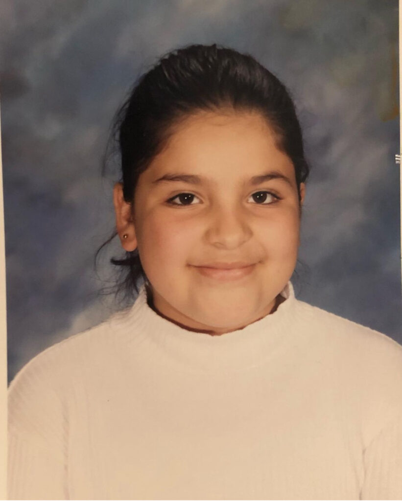 A little Mexican girl with her brown hair in a ponytail with little round gold earrings. She’s wearing a white turtleneck, looking directly at the camera. The background is a light and dark, navy and white backdrop.