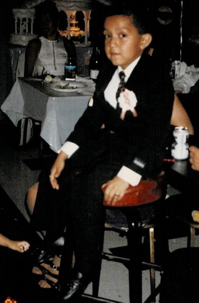 a little Mexican boy, wearing a black suit with a white shirt and a black tie with black loafers. He is sitting on a black stool with a red cushion looking directly at the camera. In the background is a large wedding cake and tables with white tablecloths. 