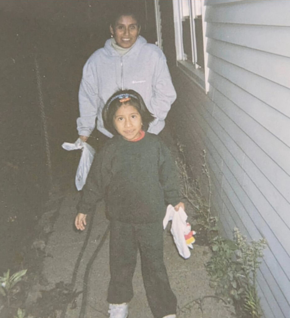 a little Mexican girl with shoulder length hair, staring directly at a camera she’s wearing a forest green sweater with matching sweatpants and she’s wearing white tennis shoes. She has a plush toy in her left hand and she’s standing outside in a little pathway with cement and bushes next to a house with white panels. Standing behind her is an adult wearing a gray hoodie with her right hand inside a pocket of her hoodie and the left hand holding a white plastic bag. She is looking directly at the camera and smiling.