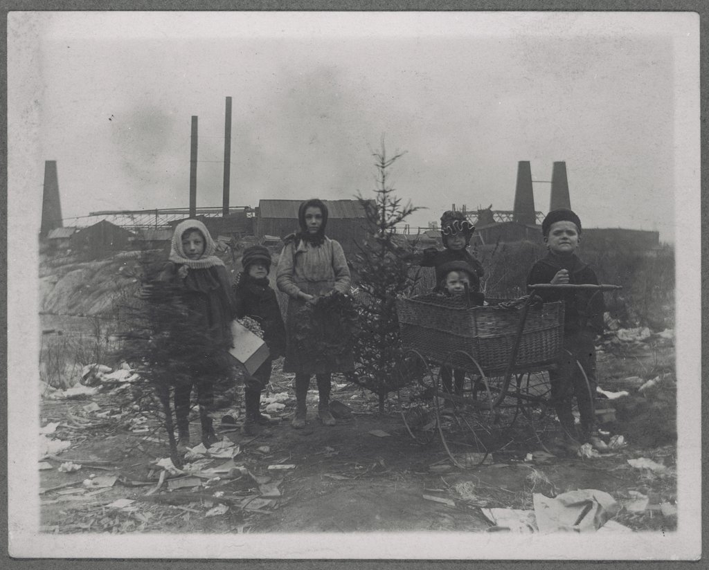 A very old black and white picture of six white children standing near an old factory with garbage around them. From left to right: A little girl holding a small tree that’s been cut at the trunk with her head covered. Next to her is a small boy holding a box. Next to the little boy is a taller girl with her head covered holding a Christmas wreath. Next to her is a tree that has been cut at the trunk and is slightly taller than she is. Holding onto the tree standing next to it is another little girl with her head covered. In front of her isn’t old baby carriage with a baby in it. And next to the carriage is a little boy with a hat holding onto the carriage.