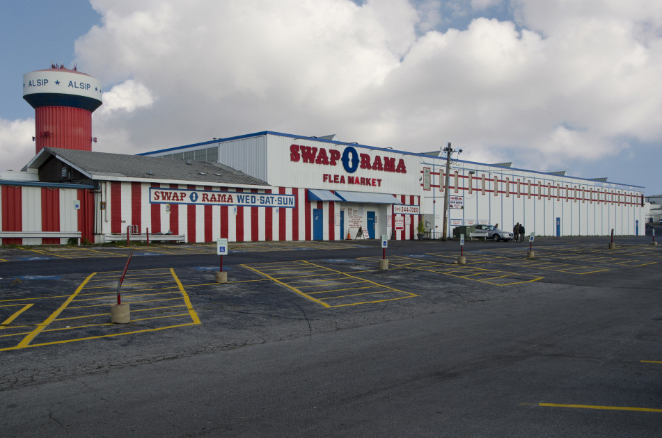 a large parking lot with black asphalt and yellow lines indicating a parking space. There are several signs indicating parking for disabled patrons. A large white building with red stripes. The front of the building has large red lettering that says Swap-O-Rama flea market. The doors to the building is blue.