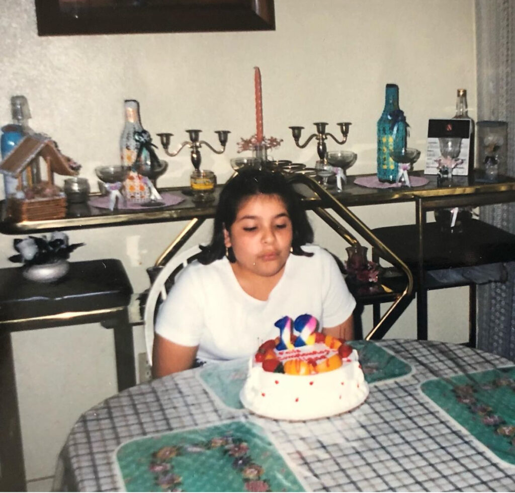 a 12 year-old girl sitting in the kitchen table with shoulder length hair and golden earrings wearing a dress that’s white at the top and navy at the bottom. She’s blowing out the candles of a cake that is all white with red dots, and has peaches and strawberries at the top. The candles on the cake are white pink, blue and yellow, and they are shaped like the number one and two. The table is covered in a white and black tablecloth and covering the tablecloth is a see-through plastic covering with green and flowery placemats. Behind the kitchen table there is a decorative glass and gold furniture that has several bottles and candleholders. 