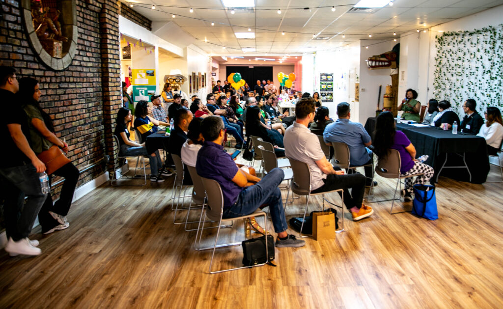  A  large gathering with people a various gender and ages are sitting in gray chairs facing a table where five people are are speaking. The there are yellow, orange, and green balloons. There is art on the wall celebrating Latin American cultures.