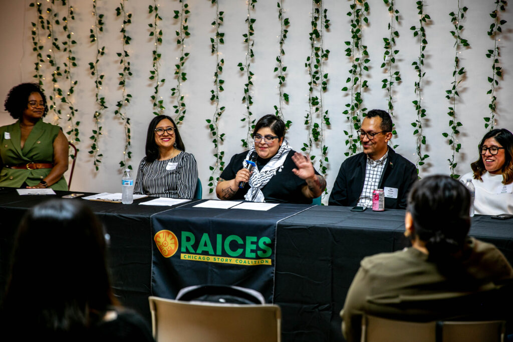 A closeup picture of five people speaking to a large crowd. From left to right and African-American woman with a short bob, wearing a green blazer and a brown belt looking towards the speakers. Next to her is a Mexican woman with brown shoulder length hair glasses, smiling with a striped black and white dress. Next to her is a short Mexican woman with black glasses, tattoos on her arms, wearing a black shirt, a black and white keffiyeh, holding a microphone in her right hand, looking down at two pieces of paper in front of her on the table. Next to her is a tall Mexican man with short curly hair glasses, a black blazer with a pin of a Palestinian flag a button up white shirt with black stripes, smiling. Next to him is a short Mexican woman with shoulder length hair glasses, smiling wearing a white sweater. They are all sitting behind a table with a black tablecloth with a yellow and orange logo, and the words rise story coalition printed on the tablecloth.