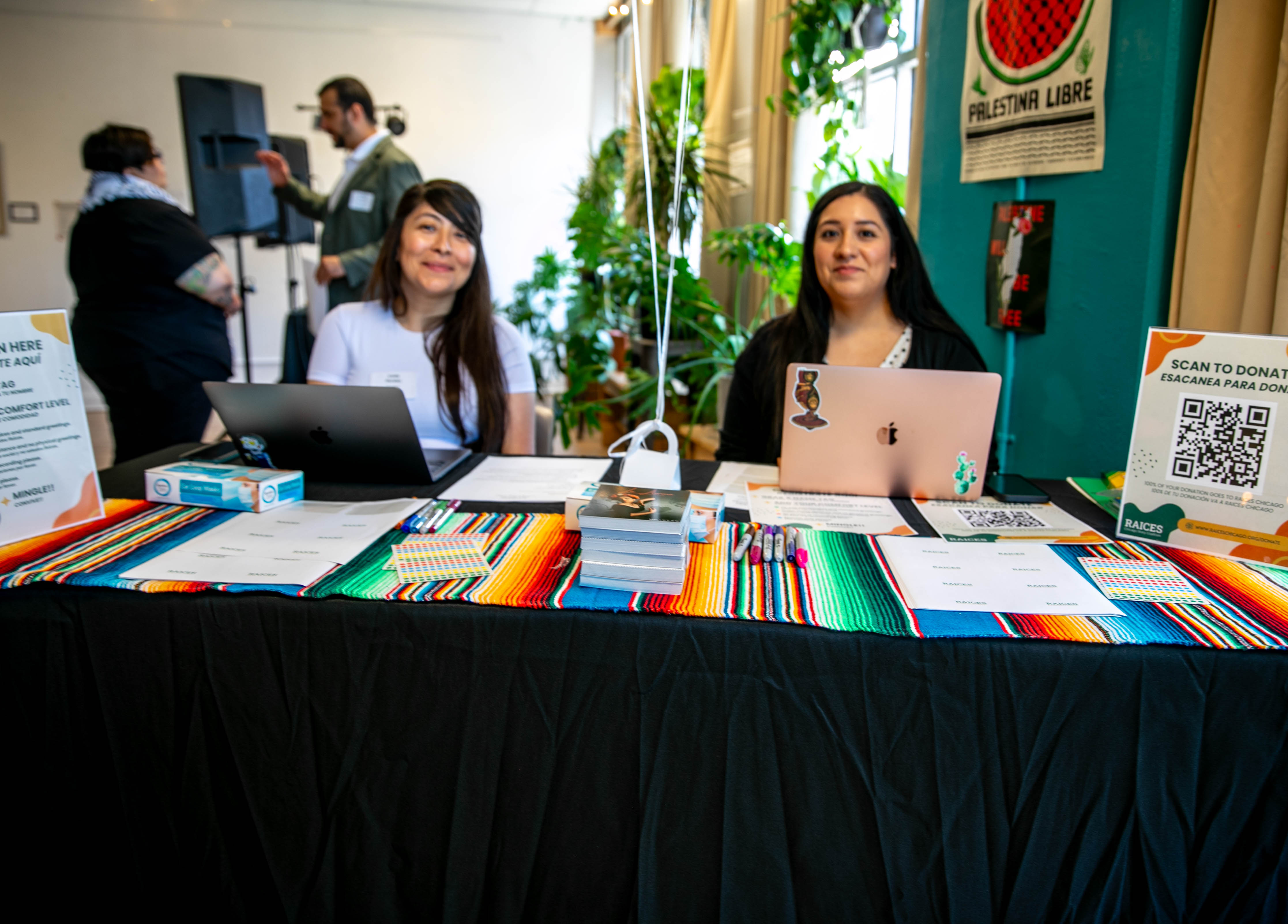 Two women sitting behind a table with a black tablecloth, a colorful serape on top with pieces of paper, face, masks, QR codes, and two laptops. From left to right: a Mexican woman with long hair and a white shirt wearing a name tag smiles looking directly at the camera. Another Mexican woman with long hair, wearing a polkadotted shirt and a black cardigan smiles directly at the camera. 