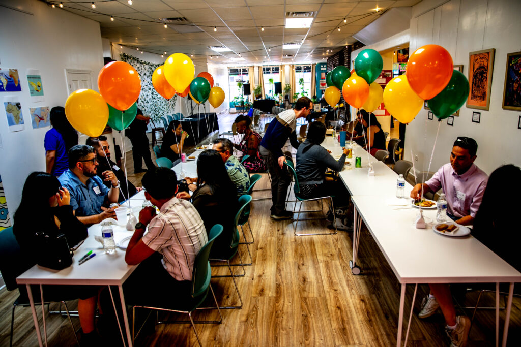 A large gathering with rows of white tables where people of various backgrounds, gender and ages are sitting eating on dark green chairs and talking. There are orange, green and yellow balloon balloons on the table tables. There are white walls with art celebrating cultural connections in Latin America. 