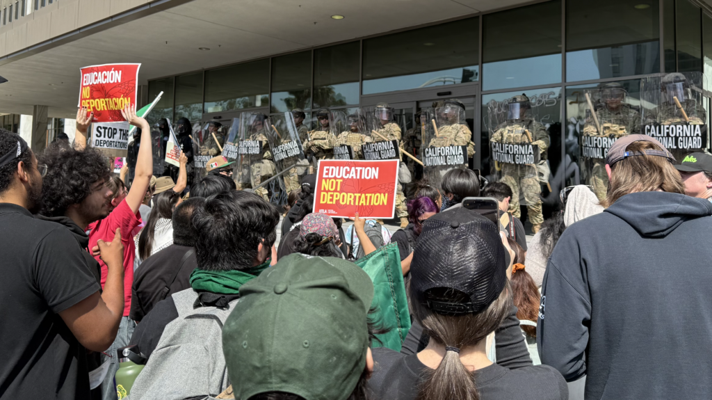 This image shows the back of protesters' heads in the foreground with some holding red protest signs. A line of men in army uniforms and plastic shields stand in front of a gray building with tall windows.