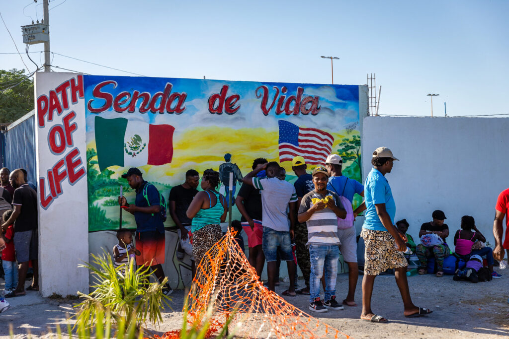 A group of twelve men, women, and children stand in front of a mural depicting a green landscape, the Mexican Flag on the middle left and the U.S. flag on the middle right. The top of the mural reads “Senda de Vida”. On the left, there is a thin, white wall with the words “Path to Life” painted in red and outlined in blue. There is a blue fence to the left with a group of men and a little boy standing. On the right side of the image, there is a white wall with a group of people sitting on the ground. People are wearing baseball caps, tank tops, tshirts, and shorts.