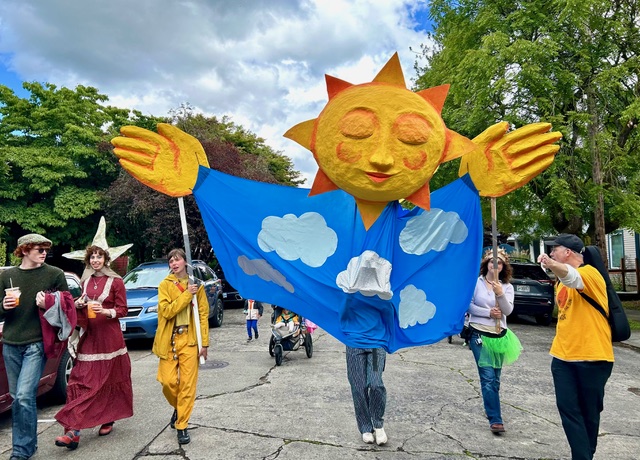 A puppet parade on the street on a sunny day. A few puppeteers maneuver a giant puppet that has the face of the sun, and its body is draped in blue clouds.