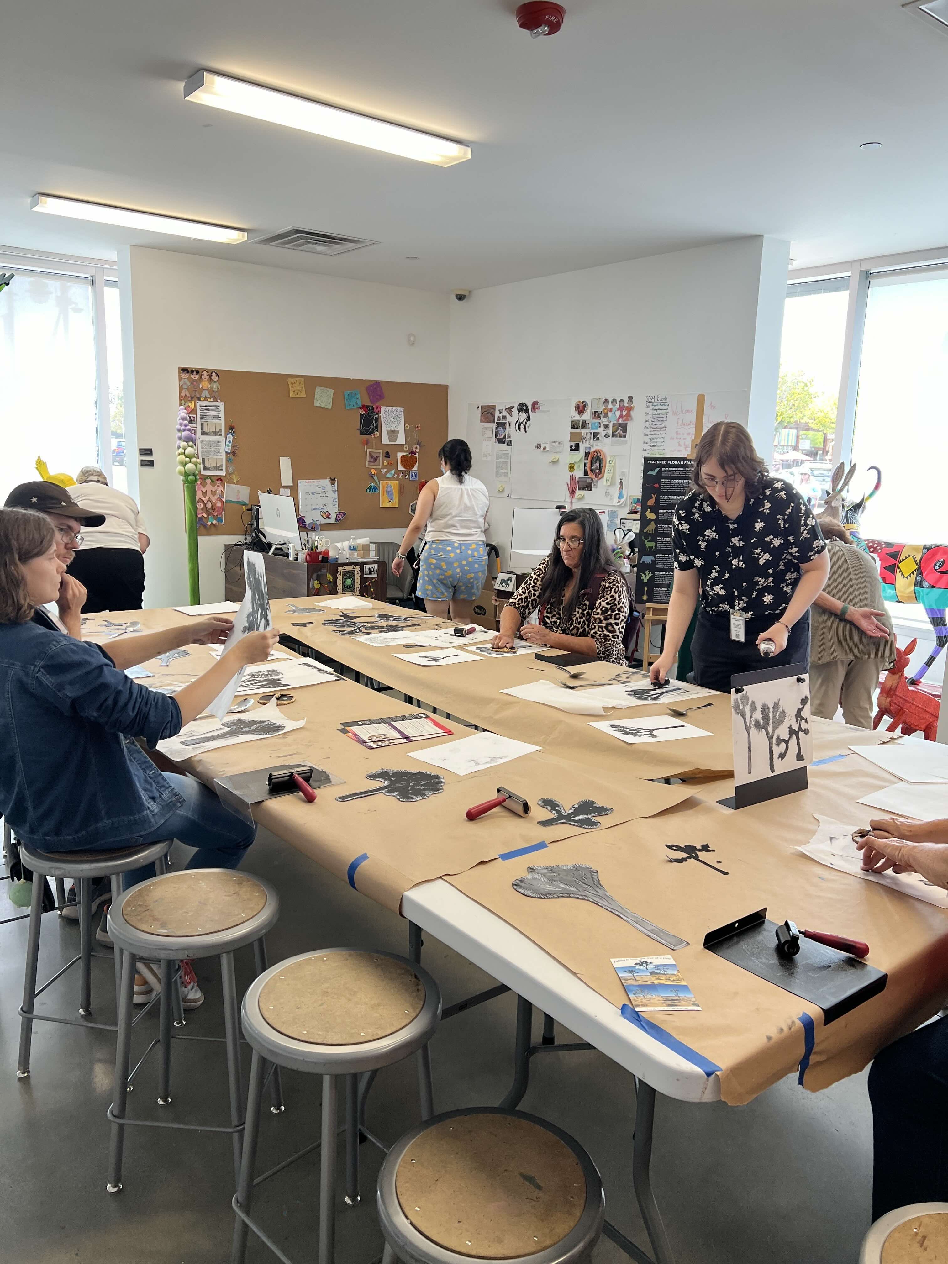 A few people are gathered around a table in an art studio. Some are looking at images, others are working on images