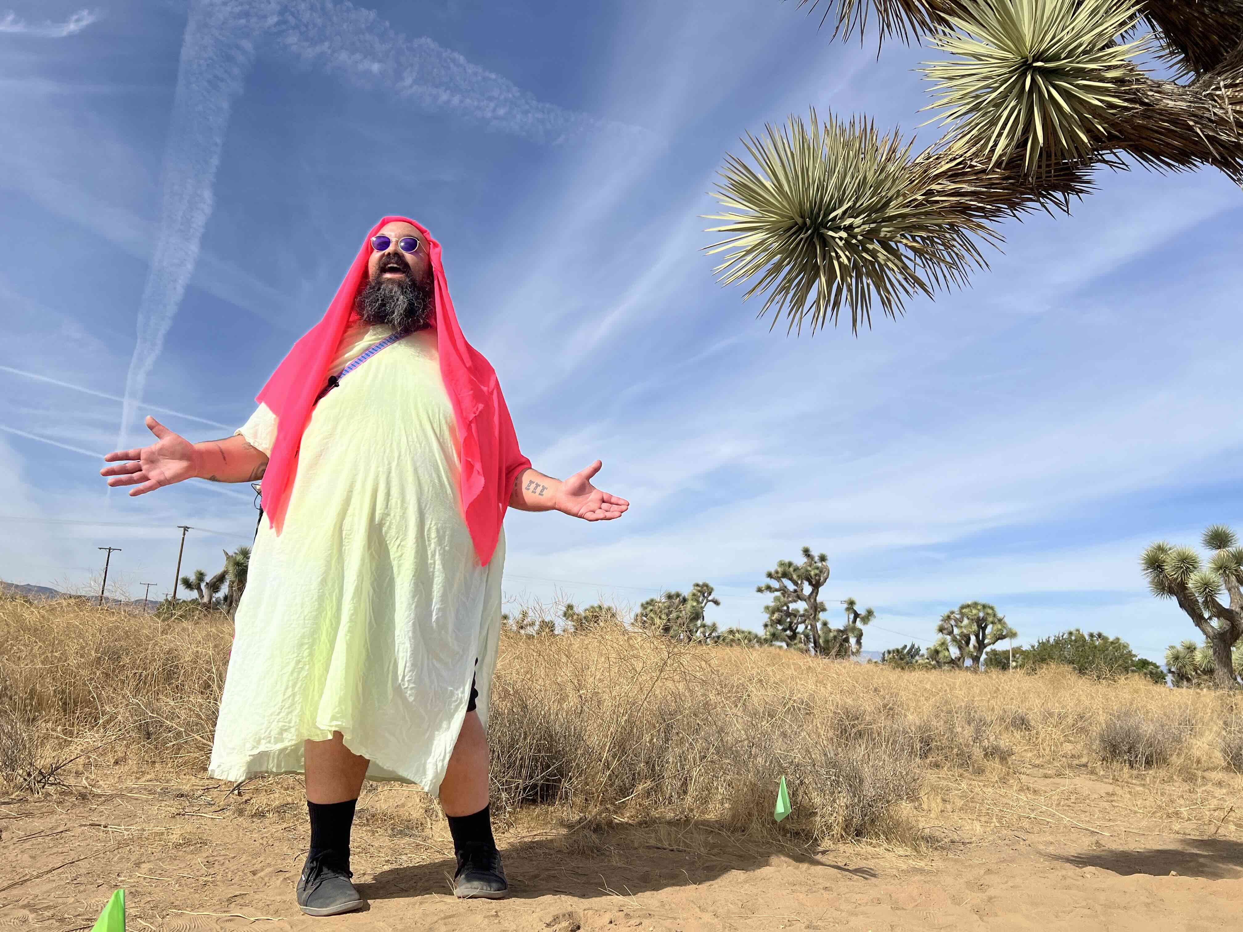 Against the expanse of the desert sky, a large man stands with his arms wide open. He is dressed in a white robe, and has a pink scarf covering his head. To the right, a Joshua Tree peeks into the frame.